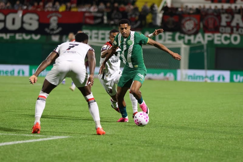 Jogador da Chapecoense, de camisa verde e branca, domina a bola sob marcação de dois adversários, em jogo na Arena Condá