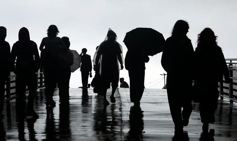 Pessoas em silhueta caminham sob chuva, algumas com guarda-chuva e capa de chuva, em passarela molhada