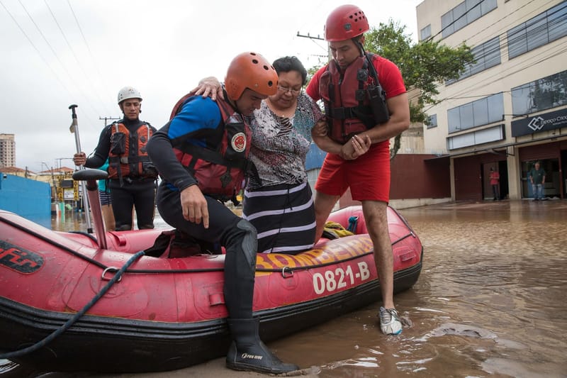Bombeiros de colete e capacete ajudam uma moradora a descer de um bote em rua alagada, com prédios ao fundo