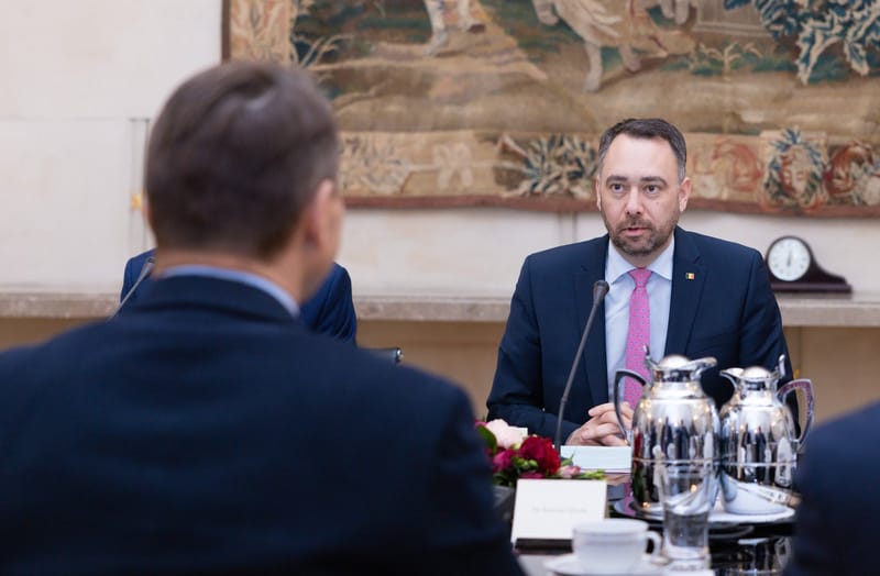 Maxime Prévot, de terno azul-marinho e gravata rosa, fala sentado a uma mesa de reunião, diante de um interlocutor de costas