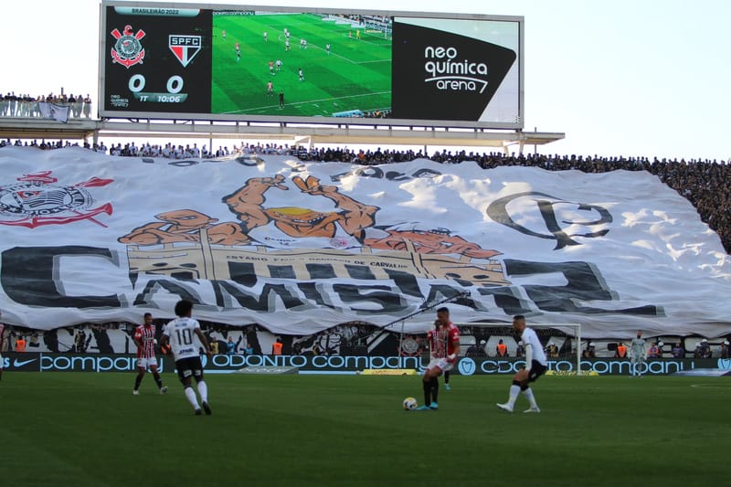 Torcida do Corinthians estende um bandeirão com o escudo do clube na arquibancada, com jogadores em campo