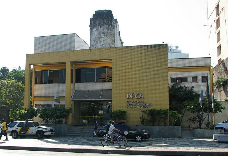 Fachada amarela de dois andares de uma delegacia de proteção à criança e ao adolescente, com viaturas estacionadas na calçada