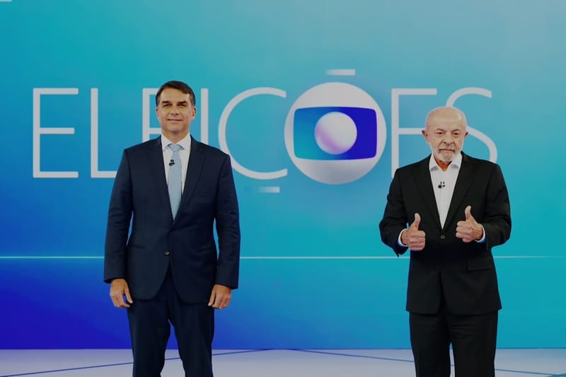 Flávio Bolsonaro, de terno azul-marinho, e Lula, de terno escuro e com os polegares levantados, lado a lado diante do painel Eleições da TV Globo