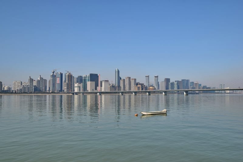 Prédios do centro financeiro de Hangzhou, na China, refletidos no rio sob céu azul, com um barco pequeno no primeiro plano