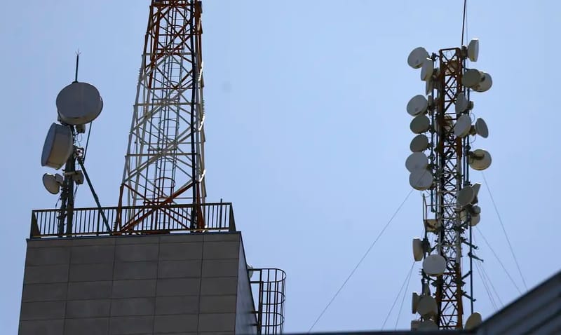 Duas torres metálicas cheias de antenas de telefonia celular no alto de prédios, contra o céu azul