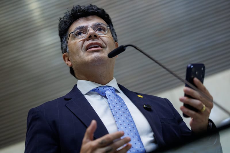Marcelo Aguiar, de óculos e gravata azul, fala no microfone da tribuna com um celular na mão