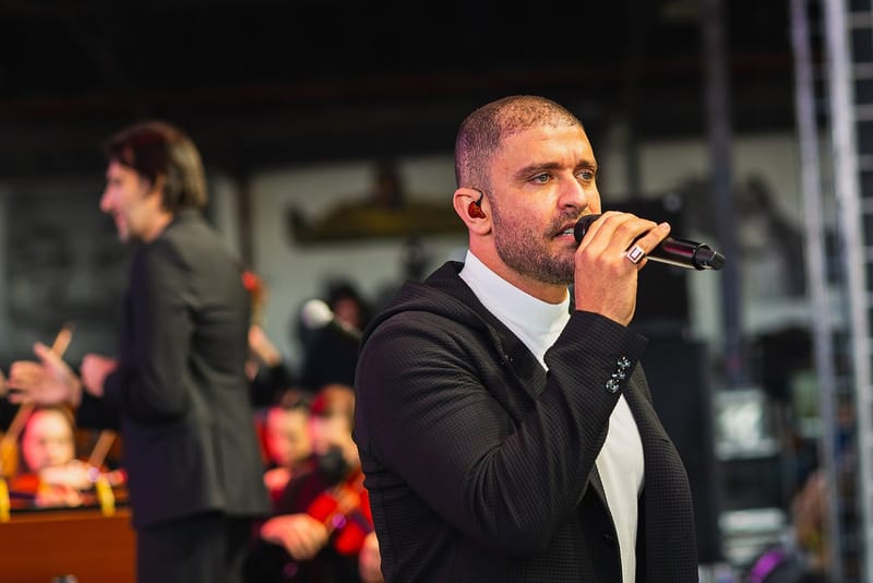 O cantor Diogo Nogueira, de blazer preto e gola alta branca, canta ao microfone em um palco, com músicos de orquestra ao fundo