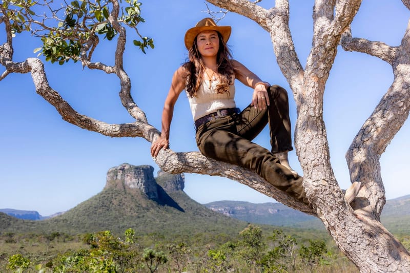 Dira Paes, de chapéu de vaqueira, sentada no galho de uma árvore, com o Morro do Pai Inácio e o céu azul ao fundo