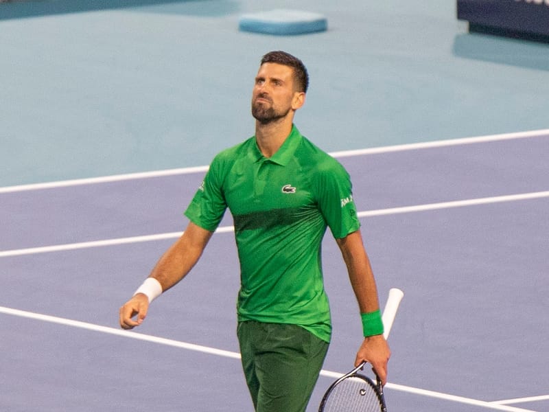 Novak Djokovic, de camisa polo e bermuda verdes, caminha pela quadra azul olhando para cima, com a raquete na mão esquerda