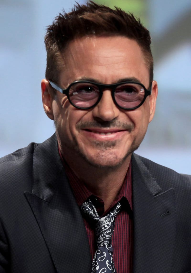 Retrato de Robert Downey Jr. sorrindo, de óculos redondos escuros e terno, durante um painel de evento