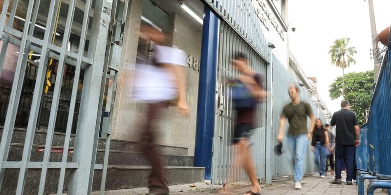 Estudantes passam em movimento diante do portão de um local de prova do Enem, em calçada ao lado de grade azul