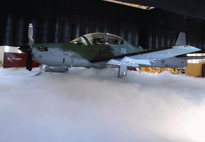 Caça A-29 Super Tucano da Força Aérea Brasileira, em tons de cinza e verde, dentro de um hangar, com neblina baixa sobre o piso