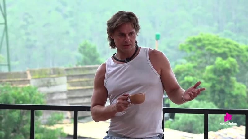 Daniel Erthal, de cabelo castanho comprido e regata branca, segura uma caneca e gesticula na varanda da sede de A Fazenda 18, com mata ao fundo