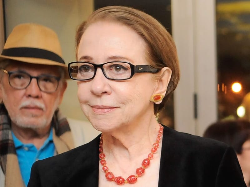 Fernanda Montenegro, de óculos de armação preta, colar de contas vermelhas e blazer preto, sorri levemente; ao fundo, um homem de chapéu