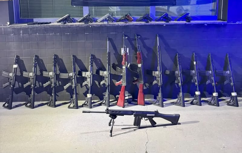 Catorze fuzis encostados em uma parede azulada e sete pistolas alinhadas no parapeito de uma janela, com um fuzil deitado no chão em primeiro plano