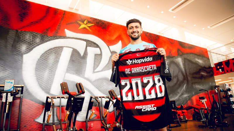 Arrascaeta sorri diante do painel com o escudo do Flamengo e segura a camisa rubro-negra com o número 2028