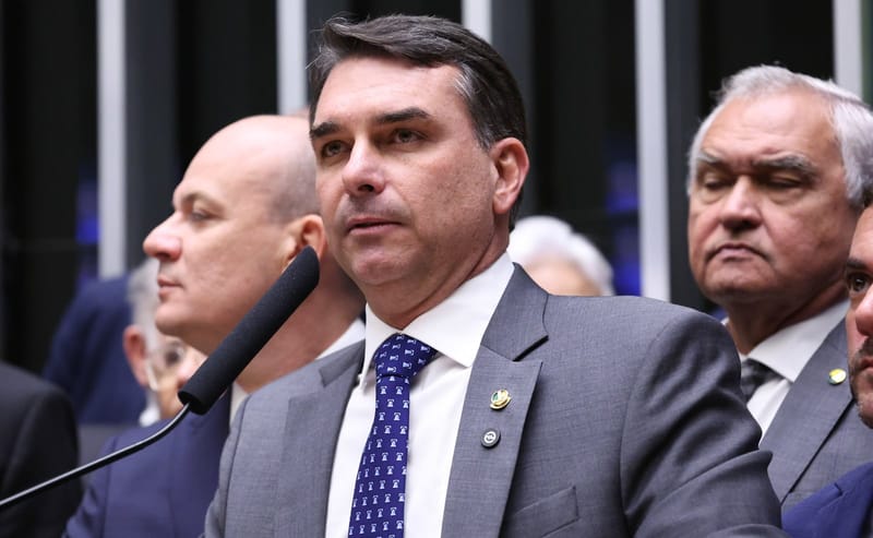 O senador Flávio Bolsonaro, de terno cinza e gravata azul, fala ao microfone no plenário da Câmara, com outros parlamentares ao fundo