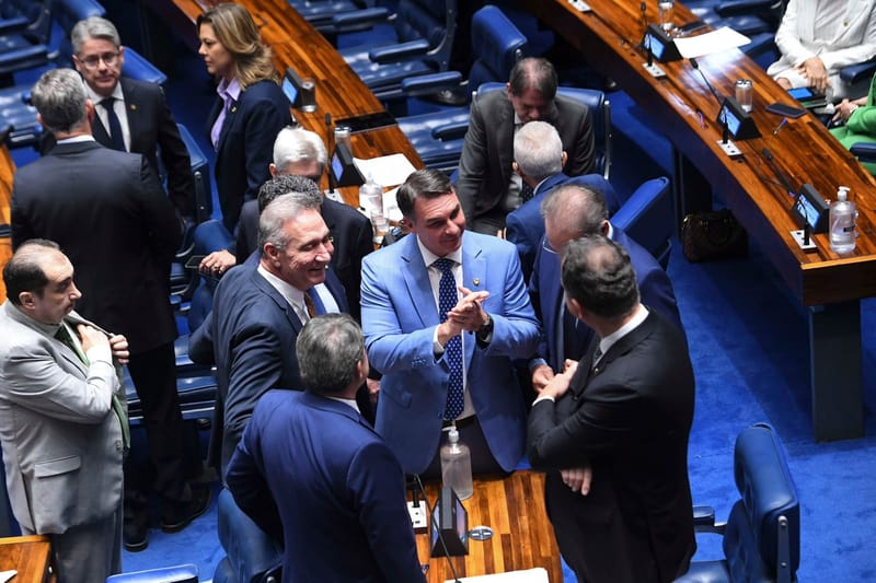 Flávio Bolsonaro, de terno azul-claro e gravata azul com bolinhas, sorri e junta as mãos cercado por senadores no plenário do Senado