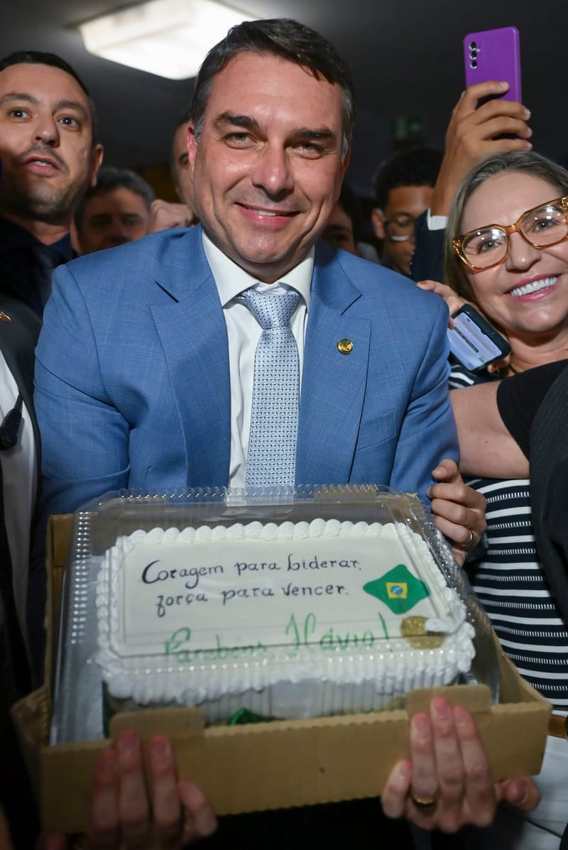 Flávio Bolsonaro, de terno azul-claro e gravata, sorri cercado de apoiadores