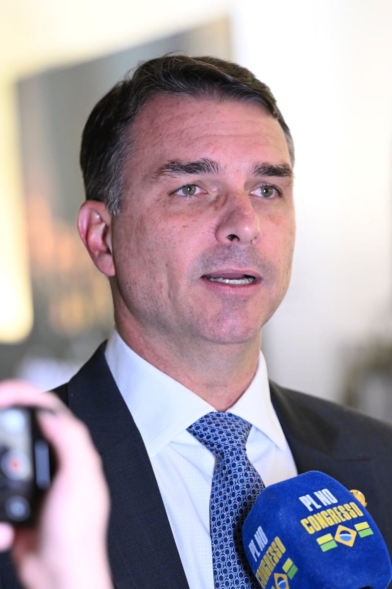 Flávio Bolsonaro, de terno escuro, gravata azul e camisa branca, fala diante de um microfone do PL no Congresso