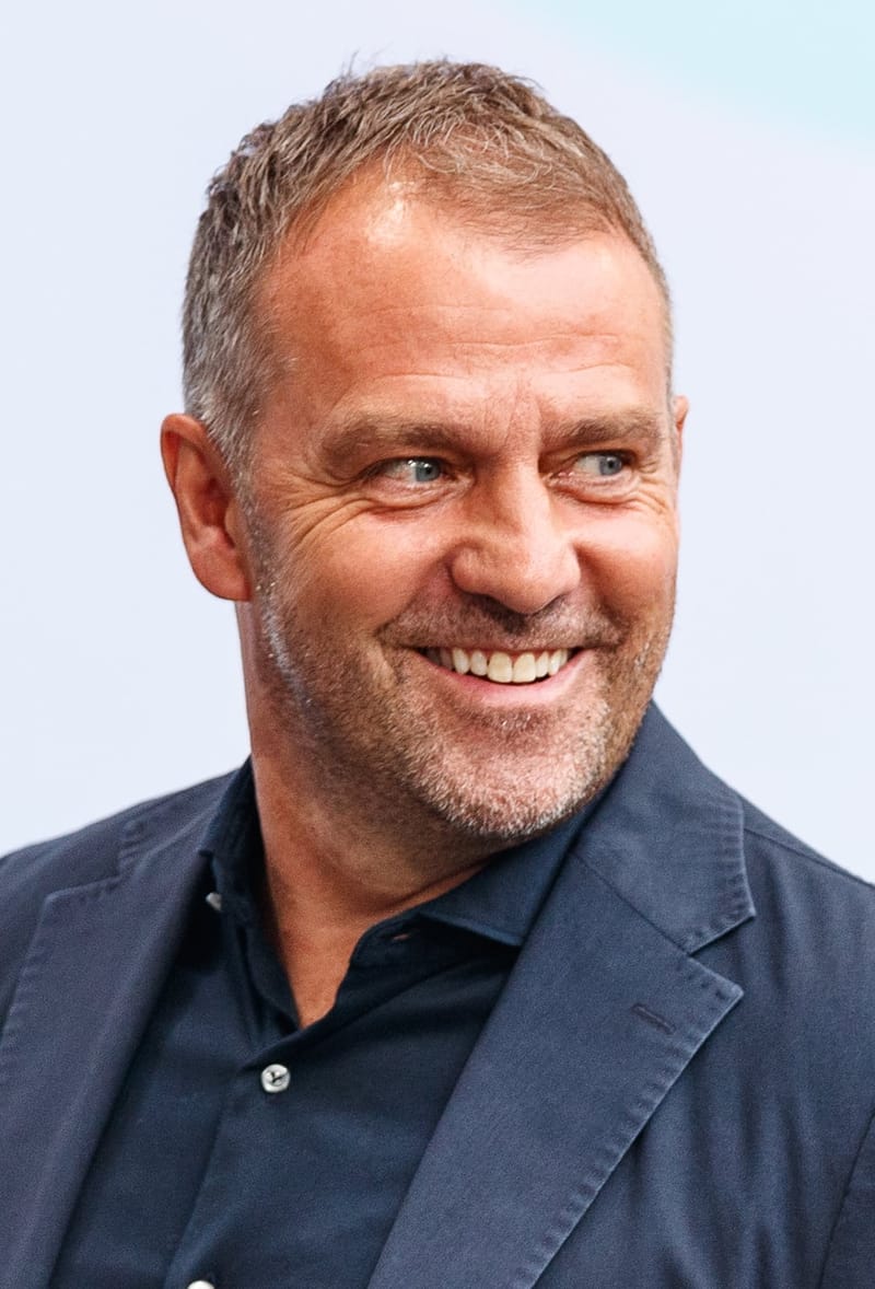 Hansi Flick, de cabelo curto e barba grisalha, sorri de lado com paletó azul-marinho sobre camisa escura, em retrato de frente
