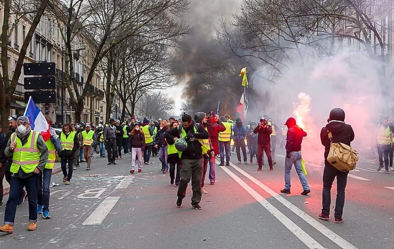 Manifestantes de coletes amarelos caminham por uma rua da França sob fumaça escura e sinalizador aceso, com bandeiras tricolores ao fundo