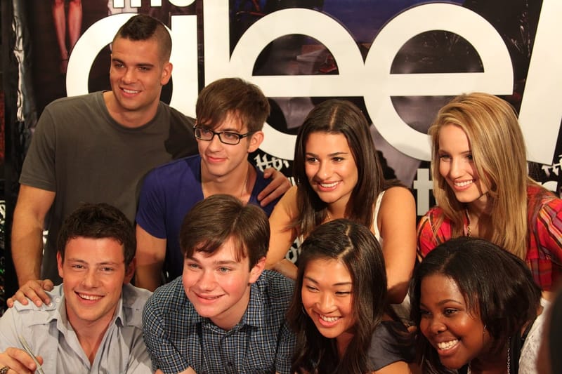 Oito integrantes do elenco de Glee posam sorrindo, lado a lado, diante de um painel com o logotipo da série