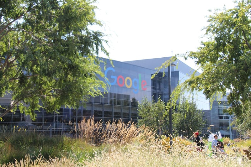 Fachada de vidro com o logotipo colorido do Google, vista entre árvores e capim, na sede da empresa em Mountain View