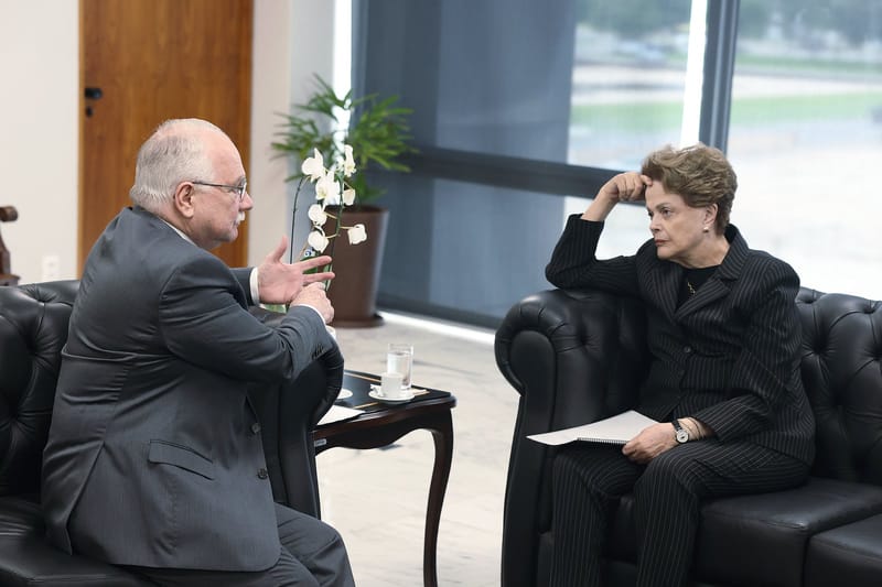 Dilma Rousseff, de terno escuro listrado e mão na testa, ouve Edson Fachin em sofá de couro preto no gabinete do STF
