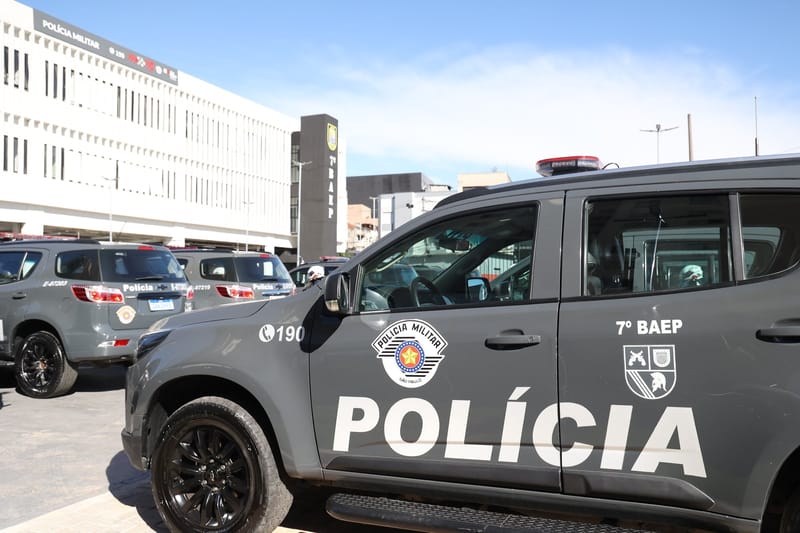 Viatura cinza da Polícia Militar de São Paulo, com a inscrição Polícia e o brasão da PM na porta, estacionada ao lado de outras viaturas diante de um batalhão