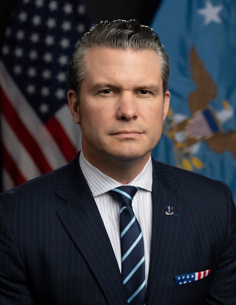 Pete Hegseth, de terno risca de giz azul-marinho e gravata listrada, posa diante das bandeiras dos EUA e do Departamento de Defesa