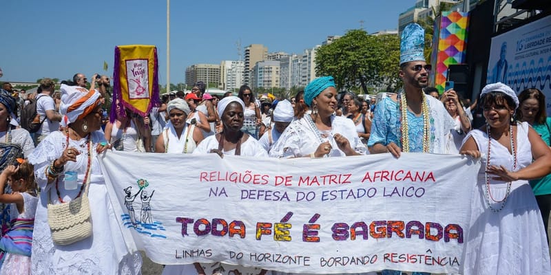 Pessoas de religiões de matriz africana, vestidas de branco e com turbantes, seguram uma faixa em defesa do Estado laico durante caminhada