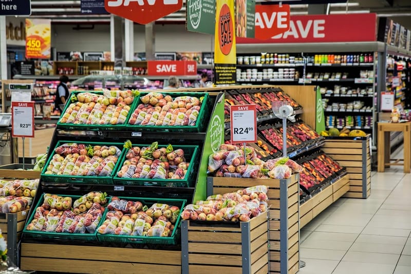Corredor de supermercado com bancas de frutas e legumes em caixas verdes e etiquetas de preço