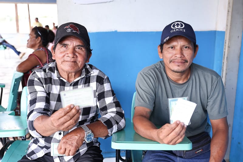 Dois eleitores indígenas sentados em carteiras escolares seguram o título de eleitor diante de parede azul e branca