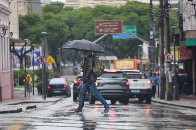 Homem de jaqueta e mochila atravessa a faixa com guarda-chuva preto em rua molhada, com carros e árvores ao fundo