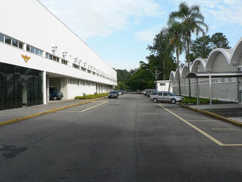 Rua do campus do ITA com prédio branco à esquerda, palmeiras e carros estacionados à direita