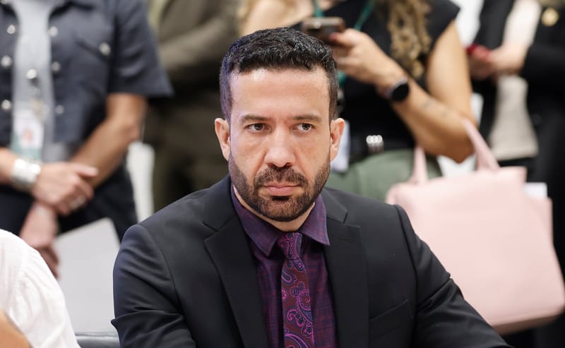 André Janones, de terno escuro e gravata roxa estampada, sentado e olhando para a frente, com pessoas desfocadas ao fundo