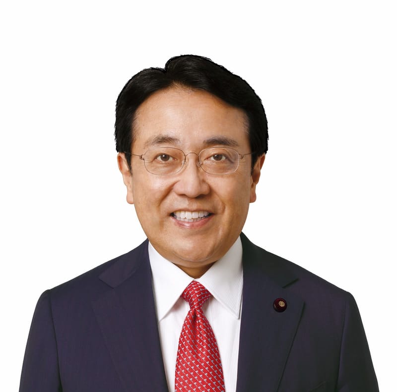Retrato de Ryosei Akazawa, homem de óculos, terno azul-escuro e gravata vermelha, sorrindo diante de fundo branco