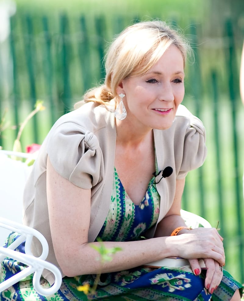 J.K. Rowling, de cabelo loiro preso e casaco bege sobre vestido estampado, sorri sentada ao ar livre