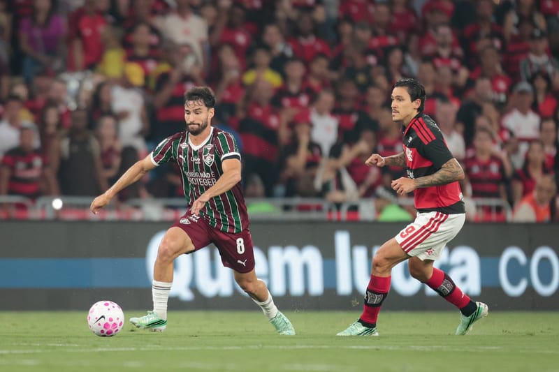 Jogador do Fluminense, de camisa grená, verde e branca, conduz a bola enquanto é marcado por atleta do Flamengo, com a torcida rubro-negra ao fundo
