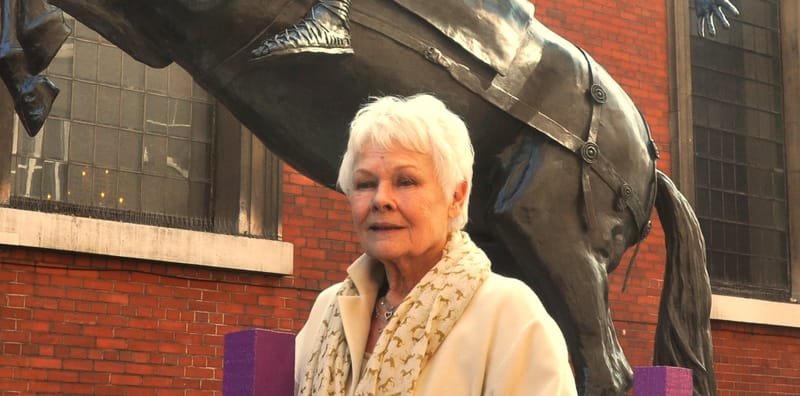 Judi Dench, de cabelo branco curto e casaco claro, em pé diante de uma grande escultura de bronze de um cavalo
