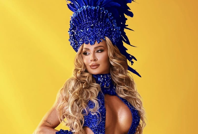 Juju Salimeni, de cabelos loiros longos, com cocar de plumas e strass azuis, em fundo amarelo