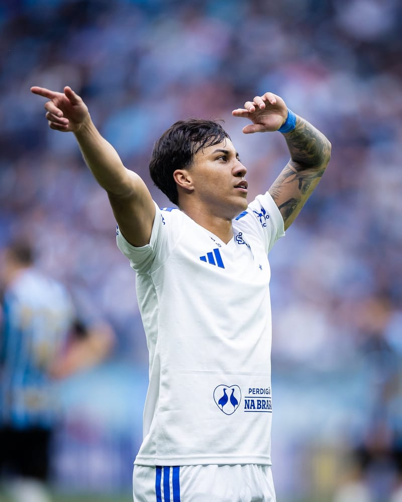 Kaio Jorge, de camisa branca do Cruzeiro, comemora de braços abertos, um deles apontando para o lado, com a arquibancada desfocada ao fundo