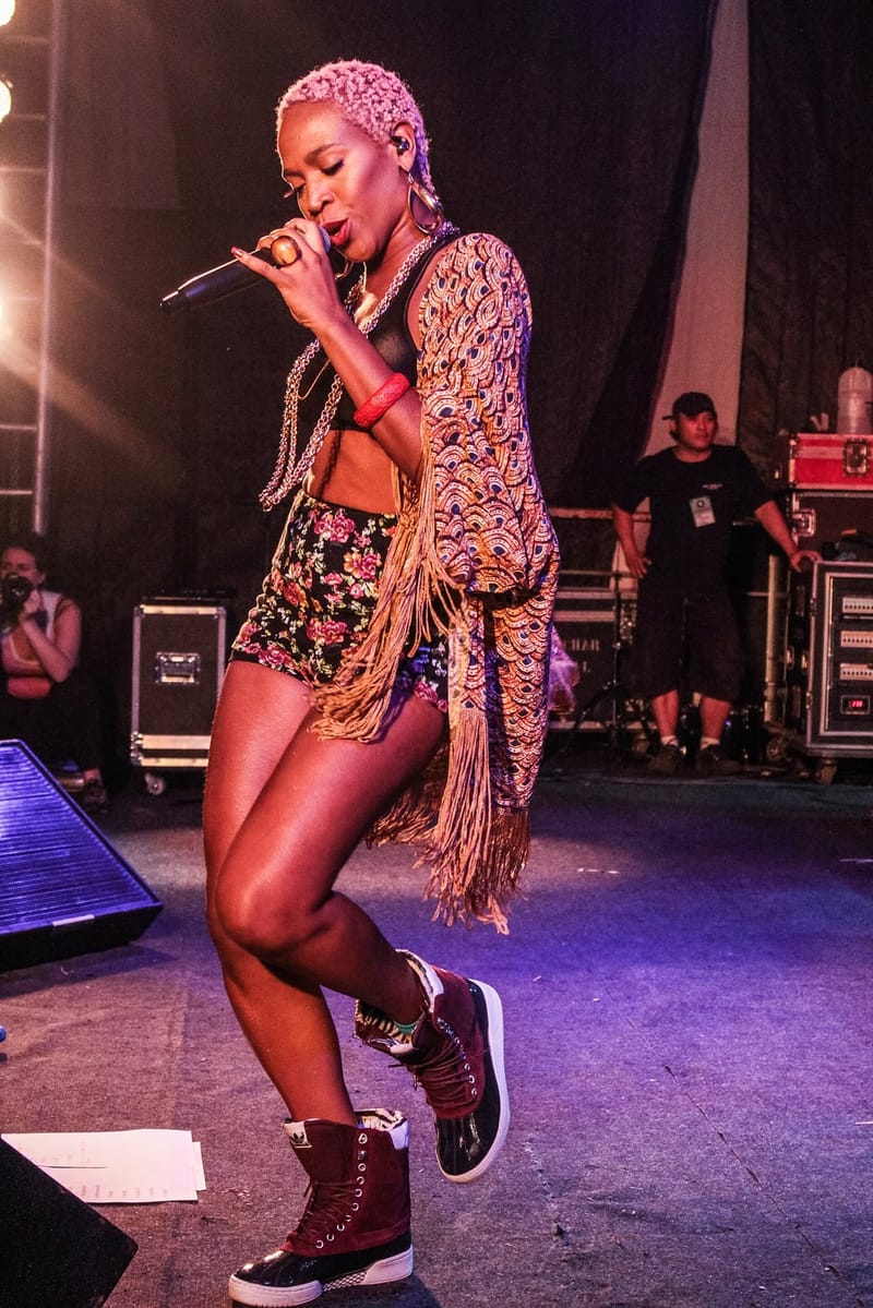Karol Conká, de cabelo curto rosado e jaqueta de franjas, canta ao microfone em um palco iluminado