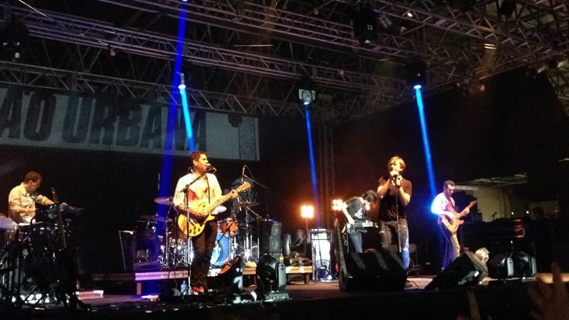 Banda se apresenta em palco com faixa da Legião Urbana ao fundo, sob feixes de luz azul