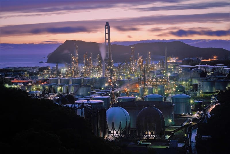 Complexo industrial com tanques e chaminés iluminados ao entardecer, à beira-mar