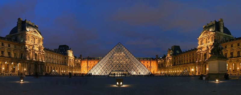 Pátio Napoleão do Museu do Louvre, em Paris, iluminado ao entardecer, com a pirâmide de vidro ao centro e as alas do palácio dos dois lados