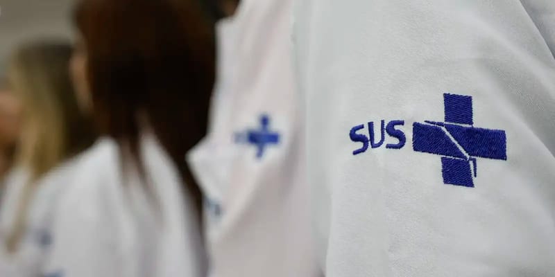 Detalhe da manga de um jaleco branco com o símbolo do SUS bordado em azul, com outros profissionais desfocados ao fundo