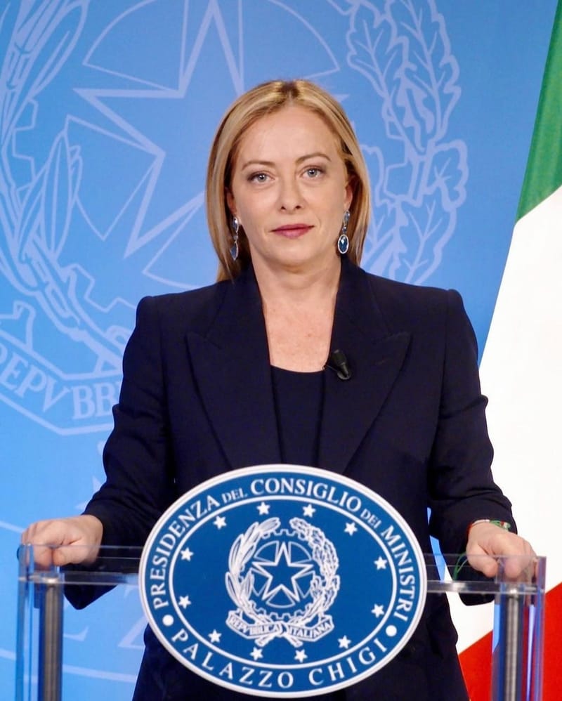 Giorgia Meloni, de terno escuro, fala atrás de púlpito de vidro com o brasão do governo, ao lado da bandeira da Itália
