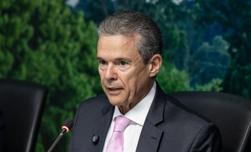 André de Paula, de terno escuro e gravata rosa, fala ao microfone diante de um painel com imagem de floresta